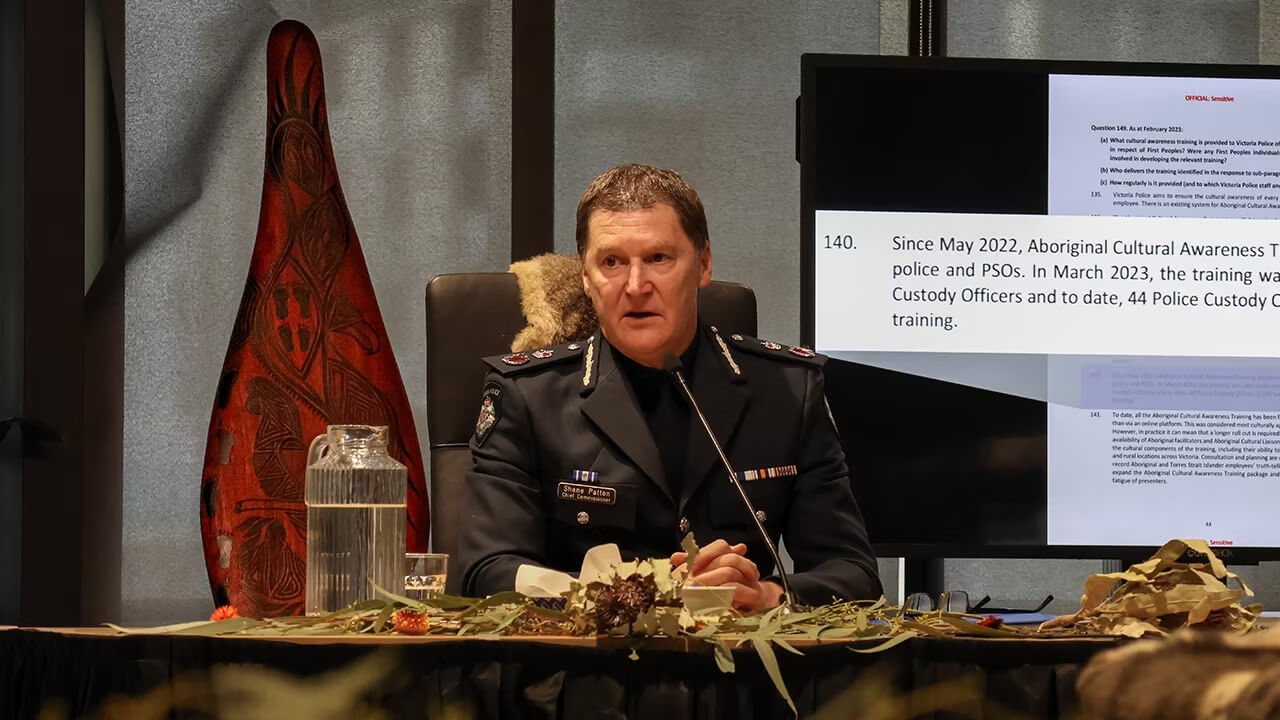 Shane Patton APM appearing at Yoorrook publics hearing in full police uniform. He is speaking into a microphone with a shield and statistics about police Cultural Awareness Training behind him.